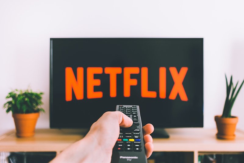 netflix in orange lettering on a tv. A remote in hand is facing the TV